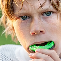 Boy with blond hair in football jersey putting green mouthguard over teeth